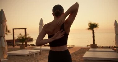 Siyah spor üniformalı esmer bir kız ellerini arkasını kolluyor ve sabahları güneşli bir plajda esneme hareketleri yapıyor..