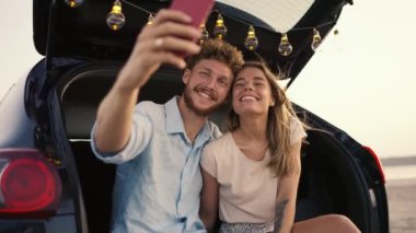 Mavi tişörtlü, kıvırcık saçlı sakallı bir adam kameraya gülümsüyor, beyaz bluzlu sarışın kız arkadaşıyla yüzünü buruşturuyor ve siyah bir arabanın bagajında komik bir selfie çekiyor.