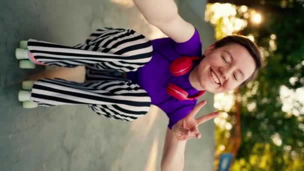 垂直视频 夏天快乐的女孩 穿着紫色上衣 戴着红色耳机 穿着条纹裤子 坐在粉色轮滑冰鞋上 坐在滑板公园里 — 图库视频影像