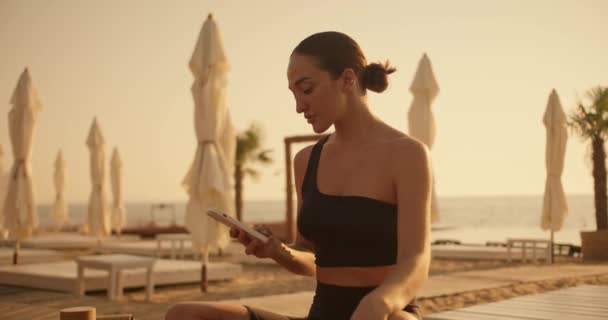一个穿着黑色上衣的黑发女孩在智能手机上工作 然后在夏天把目光投向阳光灿烂的海滩 沙滩上的黄金时间 — 图库视频影像