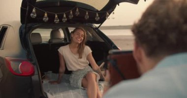 Mavi tişörtlü, kıvırcık saçlı sakallı bir adam sarışın kız arkadaşının fotoğraflarını çekiyor, kız arkadaşı gülümsüyor ve siyah arabanın bagajında Ukulele çalıyor.