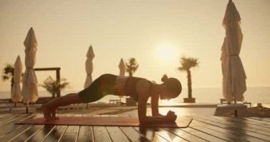 Siyah bluzlu esmer bir kız, güneşli bir plajda dirsekleriyle şınav çeker. Her tarafı tahtalarla kaplıdır. Güneş doğarken spor ve yoga.