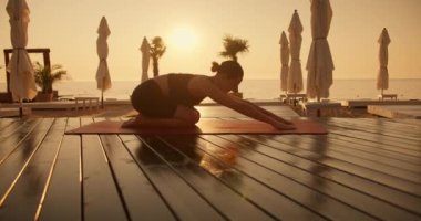 Siyah spor yaz üniformalı esmer bir kız, sarı gündoğumuyla deniz kıyısındaki kırmızı minderlerde yoga yapıyor. Kız yoga dersinde yarı kaplumbağa pozu alıyor..