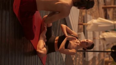 Dikey video: çıplak gövdeli esmer bir adam ve siyah bluzlu esmer bir kız meditasyon yapıyor ve ellerini siyah tahtalarla kaplı bir plajdaki kırmızı bir paspasın üzerinde namaste pozisyonuna koyuyorlar.
