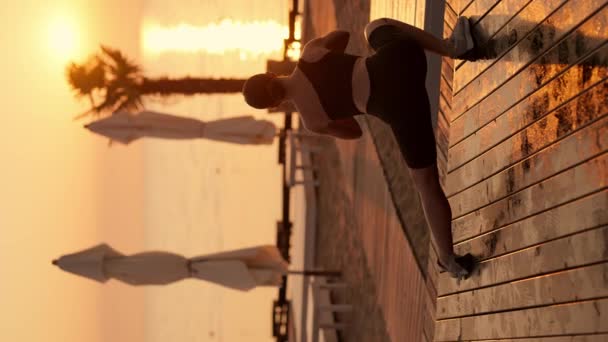 垂直录像 夏天金色日出时 穿着黑色运动服的布鲁内特女孩在阳光普照的海滩上做冲刺和伸展动作 — 图库视频影像