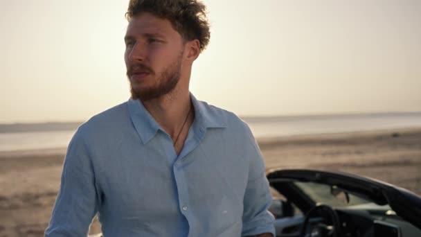 一个留着卷发 身穿蓝色衬衫 站在一辆黑色敞篷车旁边的大胡子男子卷起袖子 望着海滩上的黄天 — 图库视频影像