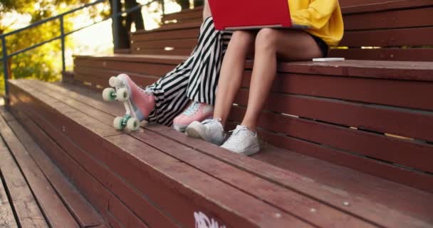 特写镜头 一个穿着条纹裤子和溜冰鞋的女孩和一个穿着短裤和白色运动鞋的女孩坐在棕色长椅上 在公园里的一台红色笔记本电脑上工作 — 图库视频影像