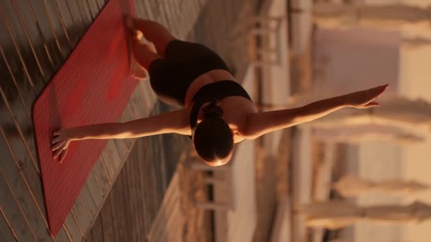 垂直视频 一个身穿黑色运动夏装的黑发女孩在一只胳膊上做着一块木板 伸出另一只胳膊的后视镜 夏季带雨伞的阳光海滩上的瑜伽和体育课 — 图库视频影像