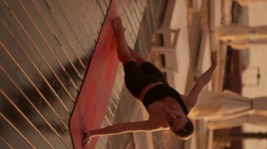 Dikey video: Siyah spor yaz üniformalı esmer bir kız bir koluna kalas takıyor ve diğeriyle güneşli bir plaja uzanıyor tahtalarla kaplı. Altın çağında yoga ve esneme dersleri