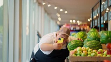Beyaz tişörtlü ve kahverengi şapkalı siyah önlüklü bir süpermarket çalışanı selfie çeker ve süpermarketteki yeşil karpuzların yanında ekşitme yapar..