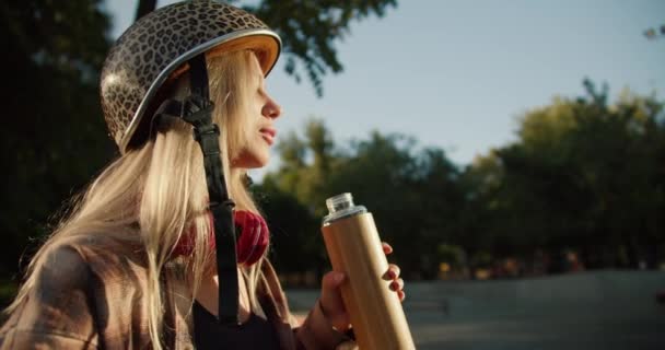 夏のスケートパークでリラックスしながら スポーツボトルから白いシャツと黒いトップの飲料水のリーパードプリントヘルメットでブロンドの女の子のクローズアップショット — ストック動画