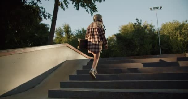 一个穿着豹纹头盔和格子花衬衫的金发姑娘 带着滑板在一个滑板公园里走上楼梯 然后开始环顾四周 把她的滑板放在旁边 — 图库视频影像
