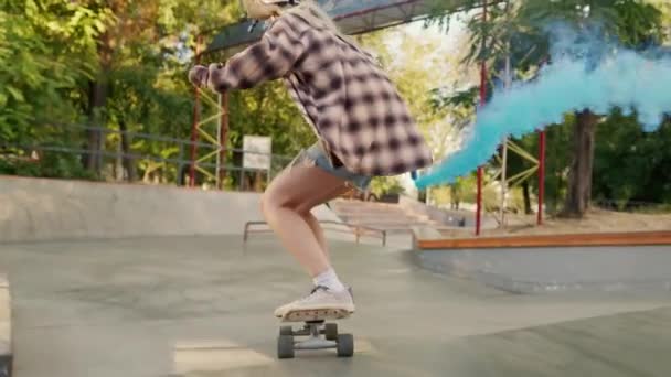 一个穿着斜纹棉布短裤和格子呢衬衫的女孩在一个滑板公园里骑着滑板 手里拿着一个烟雾炸弹 产生了蓝色的烟雾 香港一个滑雪场的浅蓝色烟花表演 — 图库视频影像