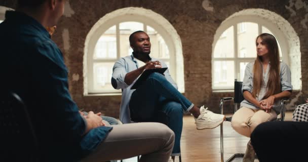 黒人男性 心理学者 ジーンズ ブリックホールで円に座っているグループセラピー参加者にインタビュー — ストック動画
