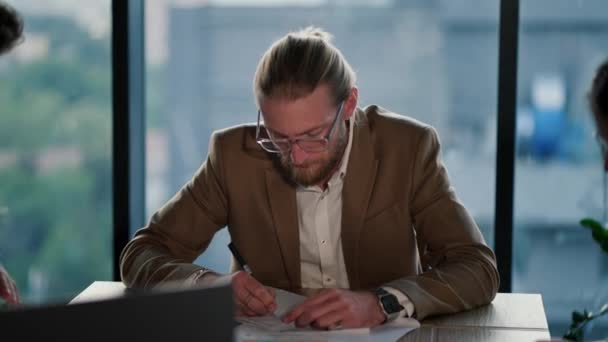 一个带着眼镜的金发年轻人 一个办公室职员 穿着浅色西服 手上拿着手表 坐在桌旁 思考问题 想想你在一个现代化的办公室里的工作 — 图库视频影像