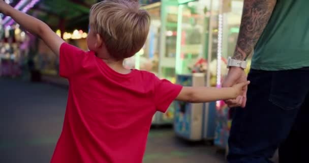 快乐的金发小儿子穿着红色T恤和他爸爸一起去看游乐园里的游乐设施 一个穿着红色T恤的小男孩和他的爸爸正在游乐园里积极地观看游乐 — 图库视频影像