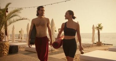 Kırmızı pantolonlu esmer bir adam ve siyah spor yaz üniformalı esmer bir kız plajda sörf tahtalarıyla yürüyor ve yoga dersinden sonra konuşuyorlar..