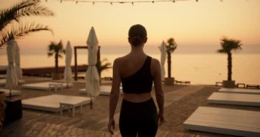 Siyah spor üniformalı, saçları tokalı bir kız sabah yoga yapmak için Golden Beach 'e gider. Güneşli bir sabah plajı, palmiye ağaçları ve güneş panelleri..