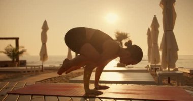 Yazın spor üniformalı esmer bir kız yoga yapar ve yazın güneşli bir sahilde kırmızı bir paspasın üzerinde kuş pozu verir. Yoga dersi sırasında akrobasi ve yoga kombinasyonu..