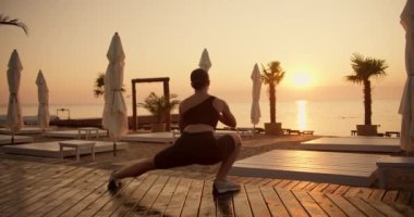 Siyah spor yaz üniformalı esmer bir kızın güneş doğarken güneşli bir plajda esneme hareketleri yapışının arka görüntüsü. Palmiye ağaçlarıyla kaplı plajda spor yapmak.