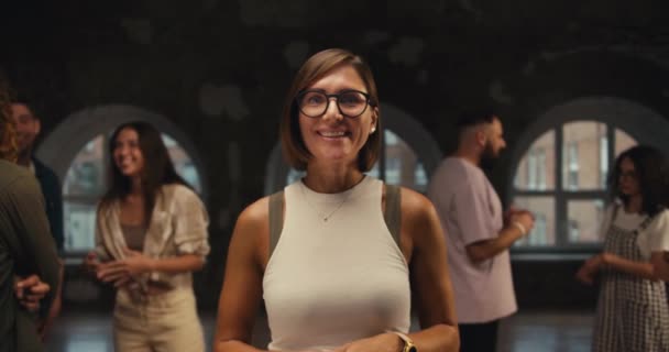 メガネのボブヘアスタイルと白いTシャツを持つ少女心理学者は レンガ造りの建物で彼女のグループ療法の背景に対してカメラを見ます — ストック動画