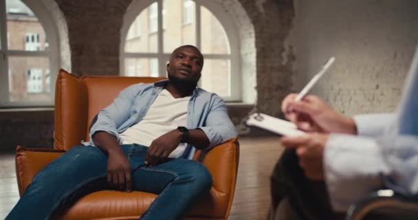 Man Black Skin Blue Shirt Jeans Sits Light Brown One — Stock Video