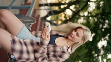 Kaykay parkında gri bir bölmede oturan ve beyaz telefonuyla sosyal ağlarda gezinen sarışın bir kızın dikey videosu. Telefonuyla mutlu sarışın kız kaykay parkında kayıyor.
