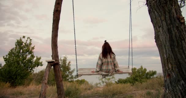 特写拍摄从后面 一个黑发女孩穿着格子衬衫在城外的木制秋千上摆动 在海边的乡间休息 — 图库视频影像