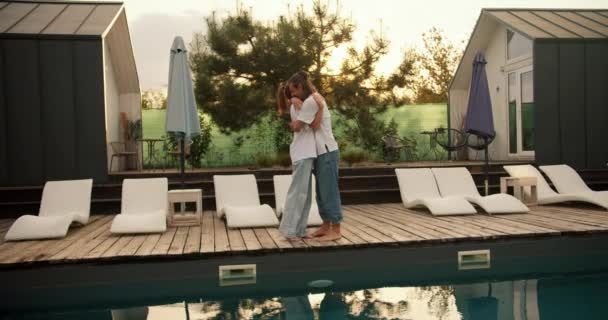 一个男人和他的女朋友穿着白色的T恤衫站在游泳池边的日光浴床旁边 在日落的背景下拥抱着 在乡间别墅休息 — 图库视频影像