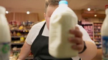 Beyaz tişörtlü, siyah önlüklü, aşırı kilolu bir erkek süpermarket çalışanı süpermarketteki bir rafta büyük plastik süt şişeleri seriyor..