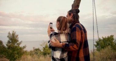 Arkasından ateş eden kareli tişörtlü bir adam, telefonuyla deniz manzarasının fotoğrafını çekerken kareli gömleğiyle kız arkadaşına sarılıp okşuyor. Kır evinde dinlen..