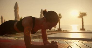 Siyah spor yazlık takım elbiseli esmer bir kız Sunrise 'daki bir plajda kırmızı bir paspas üzerinde tahta egzersizleri yapıyor. Güneşli sahil tahtalarla kaplı..