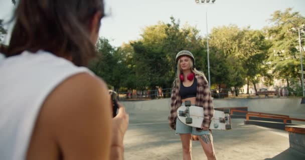 一位穿着白色T恤的黑发男子拍摄了一张照片 照片上的金发姑娘穿着格子衬衫 相机上有一块滑板 可以在夏天的一个滑板上立刻打印出一张照片 — 图库视频影像