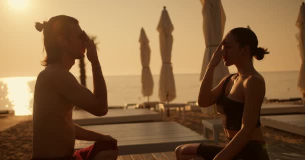 ブルネットの少女とビーチウェアのブルネットの男が瞑想をし ヨガと火の呼吸をしている 日光浴中のサニービーチでのヨガクラス — ストック動画