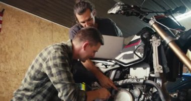 Daha deneyimli bir tamirci, öğrencisinin atölyedeki motor parçalarını düzgün bir şekilde sökmesine yardım ediyor. Garaj atölyesindeki mutlu tamirciler motosiklet teftişi ve onarımı yapıyor..