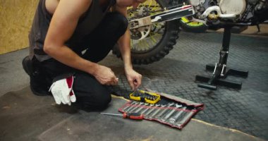 Yakın çekim: Gri tişörtlü bir adam motosikletini tamir ediyor ve teftiş ediyor. Bir tamirci ihtiyacı olan aracı tüm araçlardan seçer.