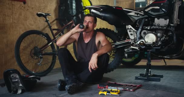 グレーのTシャツの短い髪型の男性メカニックは 彼のワークショップで彼のオートバイの近くに座っている間 暗いガラス瓶からドリンクを飲みます オートバイの修理中に休憩中に休憩する — ストック動画