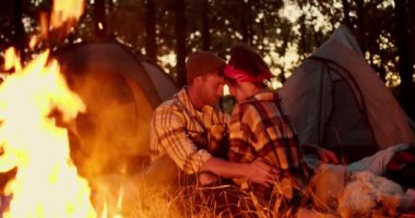 Romantik bir akşam, kareli gömlekli ve şapkalı bir adam kırmızı bandanalı ve kareli tişörtlü bir kızın yanında oturur ve çadırların arka planına karşı yakın temasta bulunurlar.