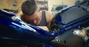 Yakın çekim: gri tişörtlü bir adam, kısa saçlı bir tamirci, atölyesinde gri motosikletini tamir ediyor - bir garaj. Atölyedeki ekipman denetimi.