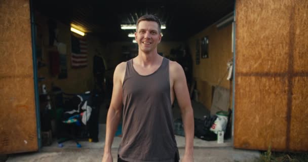 一个穿着灰色T恤 留着短发的快乐男人的画像 他把胳膊紧抱在胸前 在车库里车间前摆姿势 他最喜欢的工作是个快乐的技工 — 图库视频影像