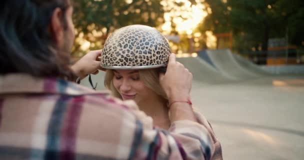 夏のスケートパークでブロンドの女の子にヒョウのプリントヘルメットを置いた平凡なシャツのブルネットの男のクローズアップショット — ストック動画