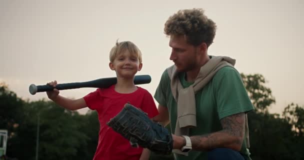 快乐的金发小男孩穿着红色T恤 手里拿着一只黑色棒球棒 穿着绿色T恤 把棒球扔到他爸爸的棒球手套里 爸爸和儿子在公园打棒球 — 图库视频影像