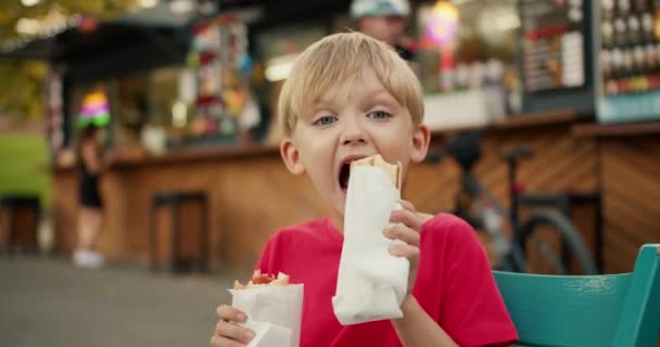 一个快乐的金发碧眼的小男孩的画像 他穿着红色T恤 一边吃着热狗 一边拿着热狗 坐在公园里一家咖啡馆的街头餐桌旁 — 图库视频影像