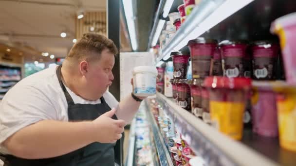 身穿白色T恤和黑色围裙的严重超重的男性超市店员在一家奶制品商店的柜台上摆放着酸奶和酸奶油 — 图库视频影像