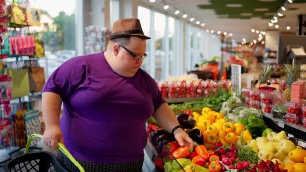 紫色のTシャツにメガネと茶色の帽子が入った太りすぎの男性は 大きなスーパーマーケットの野菜カウンターに沿って歩き トマトとズッキーニを選びます 店舗への旅のレビュー — ストック動画