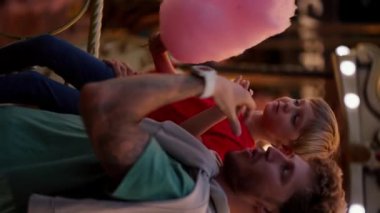 Bir babanın dikey videosu ve mavi saçlı, kırmızı tişörtlü küçük sarı oğlu akşam vakti Luna Park 'ta pembe pamuk şeker yiyorlar..