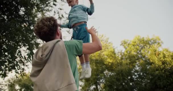 一位父亲穿着绿色T恤 卷曲的头发 把他的金发小儿子扔在公园里的一件蓝色夹克里 一个有趣的吸引小男孩 父子关系中的幸福 — 图库视频影像