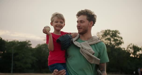 快乐的金发小男孩穿着红色T恤 手里拿着一个棒球 扔在他爸爸的手套里 穿着绿色T恤 一个男孩和他爸爸在公园打棒球的肖像 — 图库视频影像
