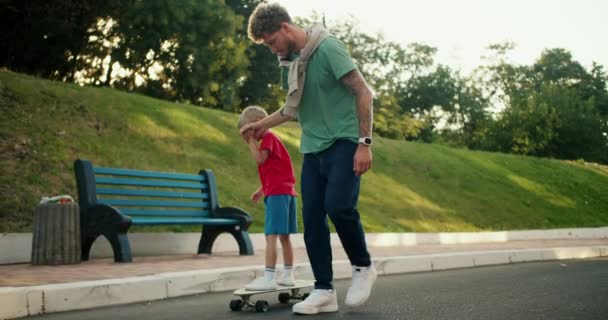 緑色のTシャツとブルージーンズの父親は 赤いTシャツと青いショートパンツの息子が公園の道路沿いのスケートボードに乗るのを助けます ハッピーパパと息子は アクティブなレジャーに時間を費やしています — ストック動画