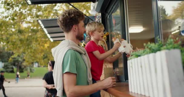 快乐的金发小男孩穿着红色T恤 坐在爸爸的怀里 一头卷曲的头发 穿着绿色T恤 从公园的一家街边咖啡馆的橱窗里收到了两个热狗 父子俩 — 图库视频影像